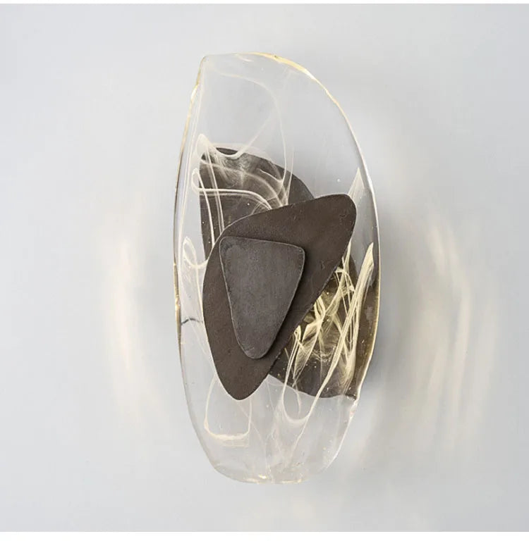 Abstract sculpture featuring dark, irregularly shaped metal pieces encased in a smooth, clear, oval resin form with white swirling patterns inside, reminiscent of the refined elegance embodied by the Luxury Crystal Wall Lamp, set against a plain light background.