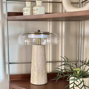The Mushroom Desk Lamp, featuring a natural stone base and glass shade, rests on a wooden shelf beside a green plant and two small labeled bottles, creating an inviting display of organic desk lighting.