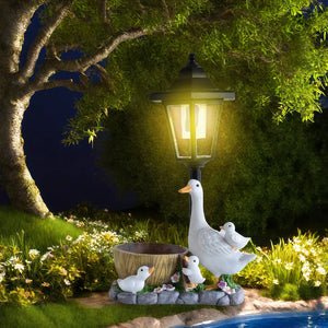 The Solar Goose Lamp, designed in the delightful shape of a goose accompanied by three gosling figurines, stands beside a small pond at night. Illuminated beneath a tree surrounded by grass and flowers, this enchanting garden decor piece is perfect for enhancing any cottage garden.