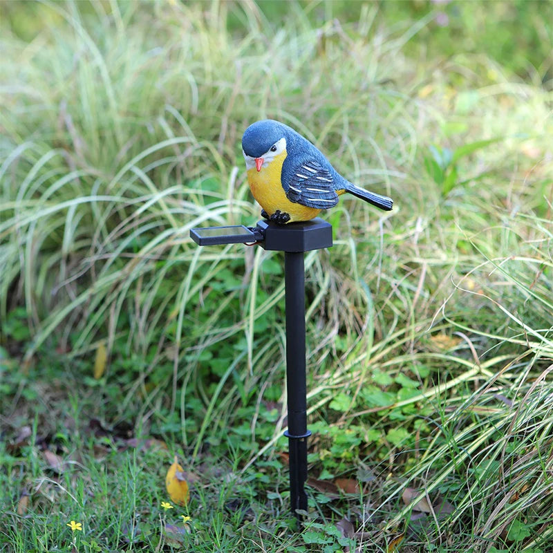 The Solar Bird Light features a decorative yellow and blue bird figurine mounted on a black garden stake, pictured set in grass amid tall green plants—making it the perfect charming songbird decor or an accent to your garden solar light collection.
