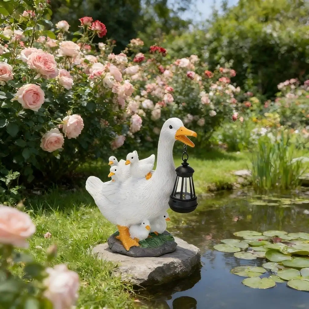 The Solar Duck Lamp, featuring a white duck carrying three ducklings and holding a solar-powered lantern, stands by a pond amidst blooming pink roses and lush green grass.