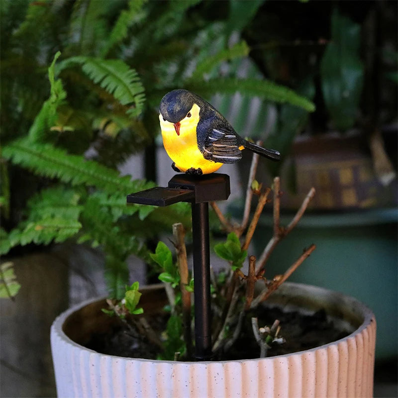 A Solar Bird Light with a yellow belly is perched on a black stand in a potted plant, surrounded by green foliage indoors.