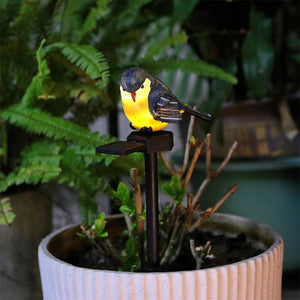 A Solar Bird Light with a yellow belly is perched on a black stand in a potted plant, surrounded by green foliage indoors.