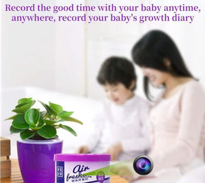 A woman and a child sit together on a bed with a green potted plant and an air freshener container placed nearby. Text above them reads: "Capture precious moments with your baby anytime, anywhere, using our Fake Decorative Display SpyCam to record your baby's growth diary.