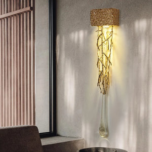 The Luxury Vertical Wall Light features a textured shade that emits warm illumination, with elegantly intertwined twisting metallic branches. Mounted on a light-colored wall near a window, it enhances any space with charm and sophistication.