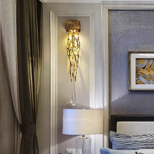 The Luxury Vertical Wall Light, featuring gold accents and a hanging crystal, beautifully complements the room's framed artwork and table lamp. Its design, reminiscent of intertwined branches, adds a touch of nature-inspired sophistication to the decor.