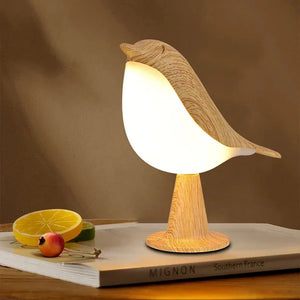 The Bird Table Lamp, featuring a modern bird-shaped design with a wooden texture, emits a warm light. It is placed on a book titled "Mignon" beside a plate with lemon slices and a small fruit. The lamp includes an intuitive touch sensor for easy control.