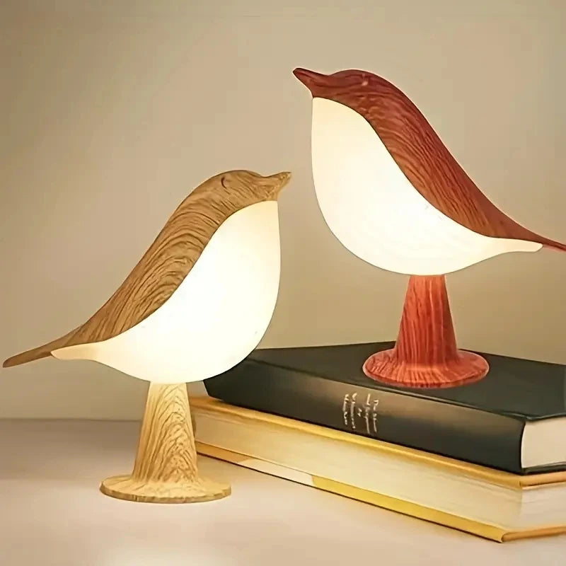 Two bird-shaped lamps from the Bird Table Lamp collection, one featuring a brown base and the other a red base, are perched atop two stacked books. These lamps emit a warm light, beautifully illuminating the surrounding area.