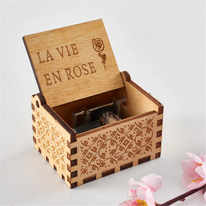 A small, handmade Wooden Music Box with intricate carvings on its sides and an open lid revealing the text "La Vie En Rose" alongside a rose engraving. Pink flowers lie beside the hand-cranked Wooden Music Box, playing classic tunes that transport you to a bygone era.