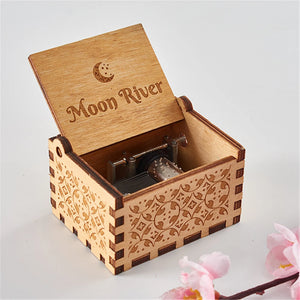 A small hand-cranked Wooden Music Box, intricately engraved with "Moon River" and featuring a crescent moon design, adorned with a flower decoration in the foreground.