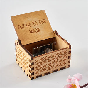 The Wooden Music Box, a small handmade piece with intricate carvings, opens to reveal its inner mechanics. The lid is propped up, displaying the words "FLY ME TO THE MOON." Nearby lies a pink flower, evoking a timeless charm reminiscent of classic tunes.