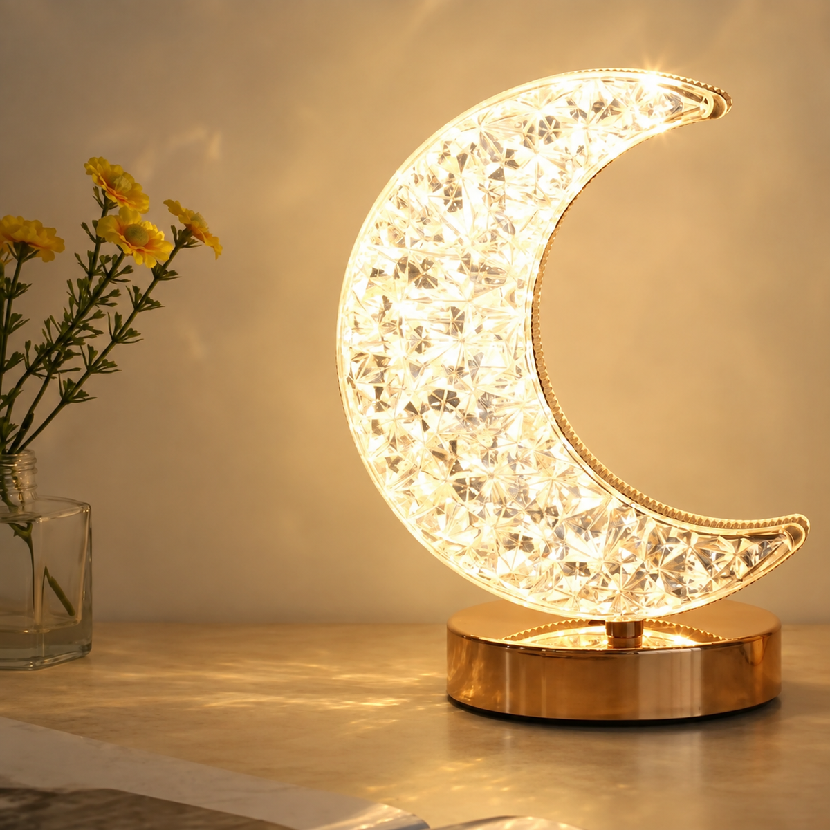 A Crystal Table Lamp with a gold base, shaped like a crescent moon, glows on a table beside a glass jar filled with yellow flowers.
