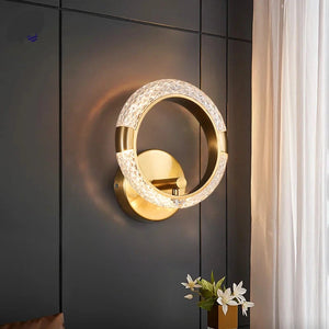 The Elegant Ring Wall Sconce, featuring a gold finish and diamond-patterned light reminiscent of a luxury wall light, encircles an inner adjustable arm. It is mounted on a dark paneled wall next to white curtains and a small vase of white flowers.