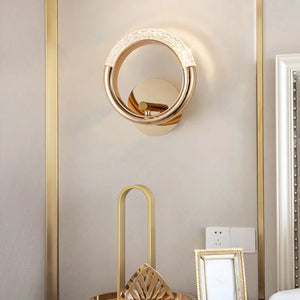 The Elegant Ring Wall Sconce, featuring a gold-toned ring design, is mounted on an off-white wall above a decorative tray, with a small rectangular frame and an electrical outlet in view.