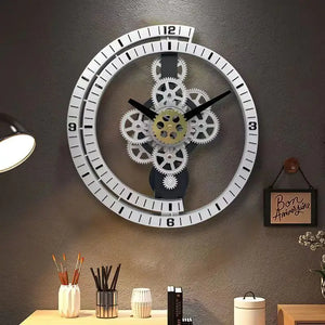 The Industrial Gear Wall Clock, showcasing its visible gears and exuding an industrial aesthetic, is mounted on a gray wall above a desk adorned with a lamp, pencils, and decorative items.