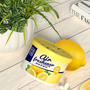 A can of air freshener with a lemon-scented label is placed on a light wooden surface next to a whole lemon and a potted plant, cleverly disguising the hidden camera inside the Fake Decorative Display SpyCam.