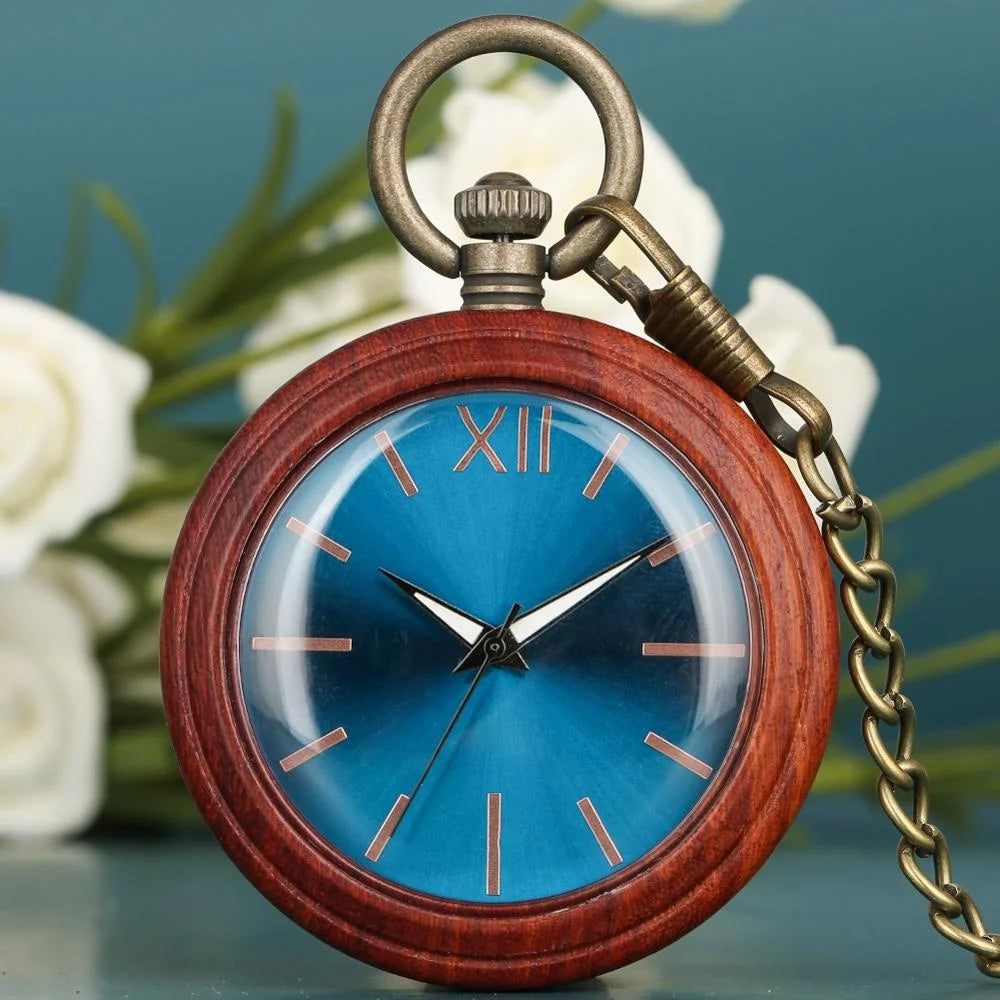 The Custom Vintage Antique Quartz Pocket Watch features a handmade wooden case, blue face, gold hour markers, and a bronze chain, shown resting against a blurred floral background.