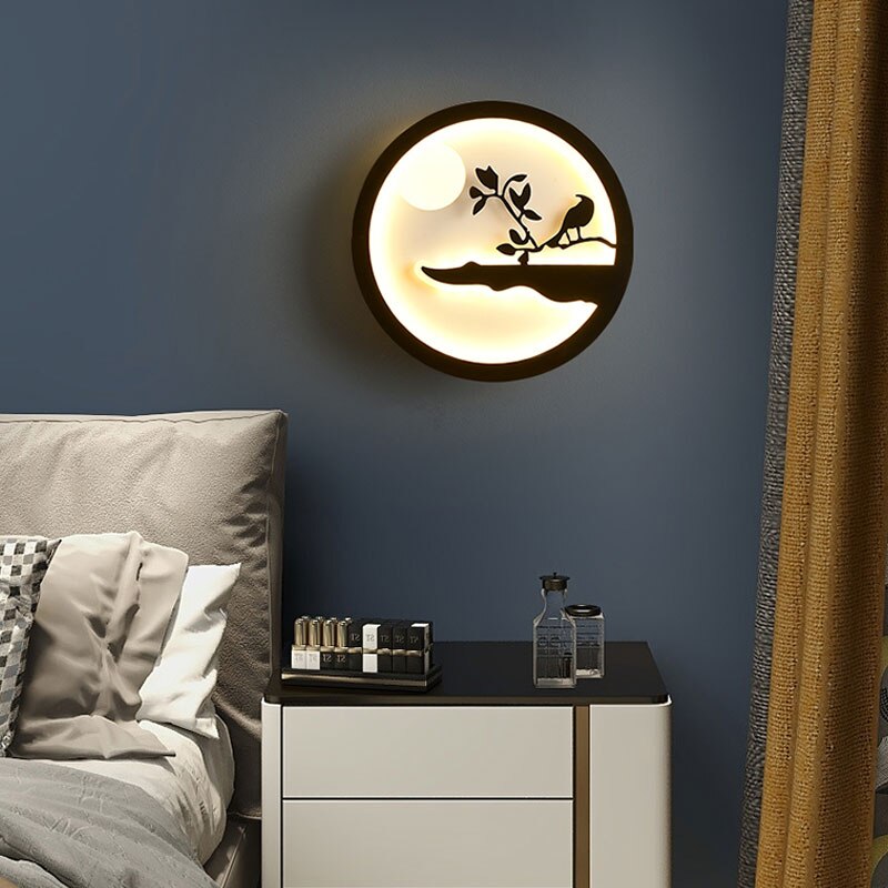 The Minimalist Wall Lamp, with its round design and bird-and-branch silhouette, emits a gentle and warm light that enhances the ambiance of a contemporary bedroom featuring a bed, cushions, and a nightstand decorated with bottles.
