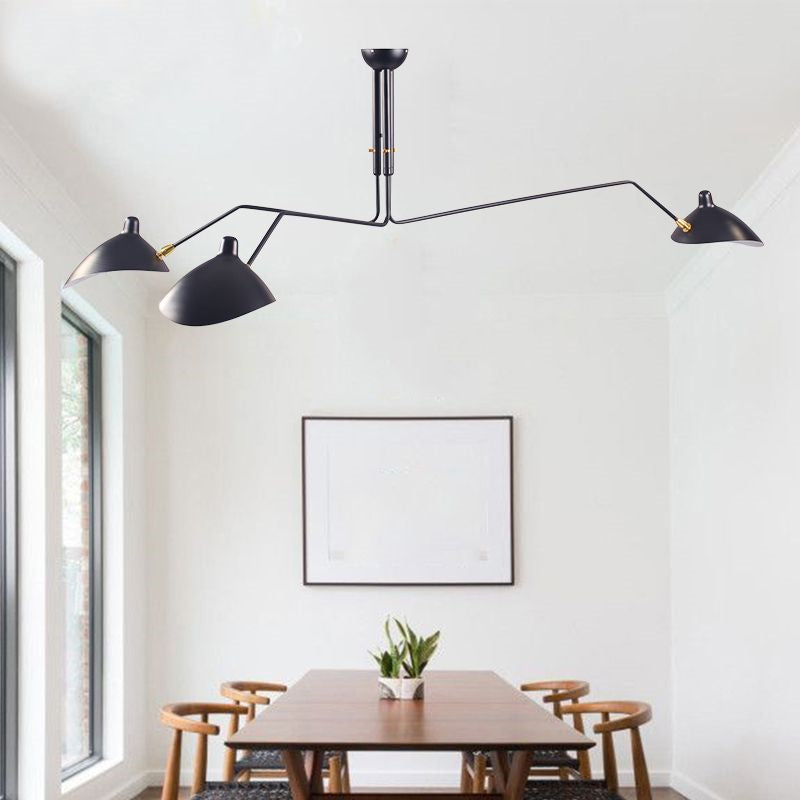 A minimalist dining room showcases a wooden table with chairs, highlighted by a Sleek Industrial Pendant Light featuring brass accents and three black arms above. A small plant decorates the table under large windows lining the left wall, with an empty white frame hanging nearby.