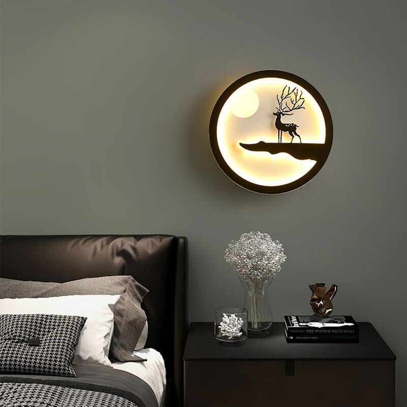 The Minimalist Wall Lamp, featuring a circular backlit design with a silhouette of a deer on a hill, emits a soft and warm glow. Elegantly mounted above a bedside table adorned with decorative items, it enhances the serene ambiance of a modern bedroom.