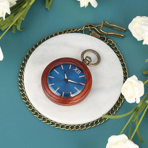 The Custom Vintage Antique Quartz Pocket Watch, a natural handmade wooden pocket watch, features a blue face with Roman numerals and rests on a white surface amid a brass chain and white flowers against a teal background.