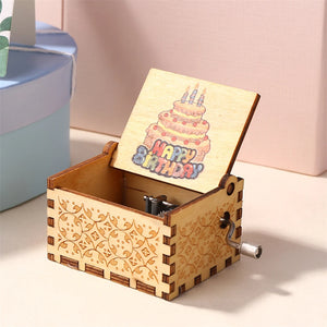 The Happy Birthday Music Box, a wooden music box with decorative carvings and a partially open lid, reveals an illustration of a birthday cake with the words "Happy Birthday." It features a hand-cranked metal winding handle on the side.