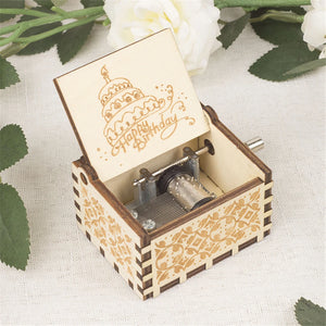 The Happy Birthday Music Box, featuring a hand-cranked mechanism and an open lid, showcases a "Happy Birthday" message along with an engraving of a cake, all surrounded by white flowers.