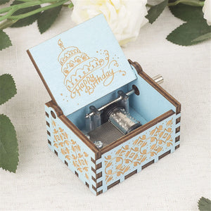 The Happy Birthday Music Box, made of blue wood and adorned with intricate designs, features an open lid that reveals a "Happy Birthday" message along with a cake illustration. It is elegantly placed on a white cloth next to green leaves and a white flower. This charming gift is perfect for celebrating special moments.