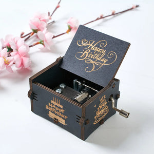 A partially opened Happy Birthday Music Box, featuring a black wooden exterior and an inscription reading "Happy Birthday" on its lid and front, revealing its intricate hand-cranked metal mechanism against a backdrop of pink cherry blossoms.