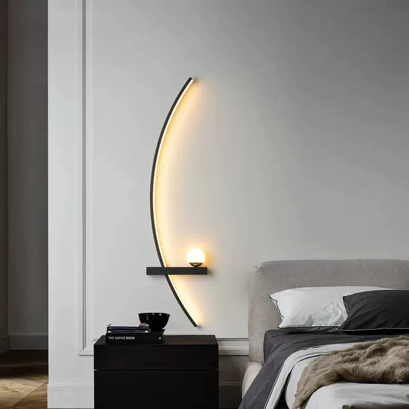 A modern bedroom features the Arc Wall Sconce, a small shelf with a round lamp, a black nightstand with books and a bowl, and a neatly made bed with a gray blanket and white pillows.