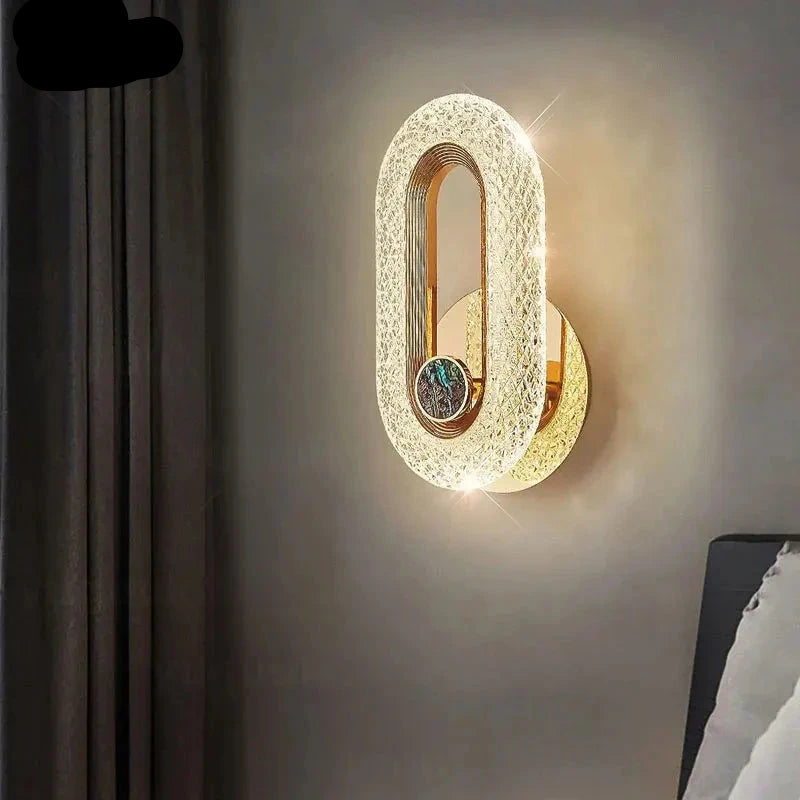 The Luxury Wall Sconce, an oval-shaped, wall-mounted LED light with an intricate design, emits a warm glow and is attached to a grey wall beside dark curtains, with a partial view of a bed.