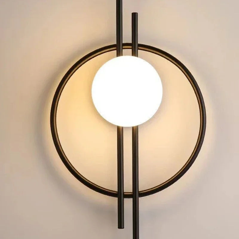 The Circular Wall Sconce with Dual Vertical Rods features a contemporary design with a glowing round bulb centered between two vertical black rods and encircled by a sleek black ring.