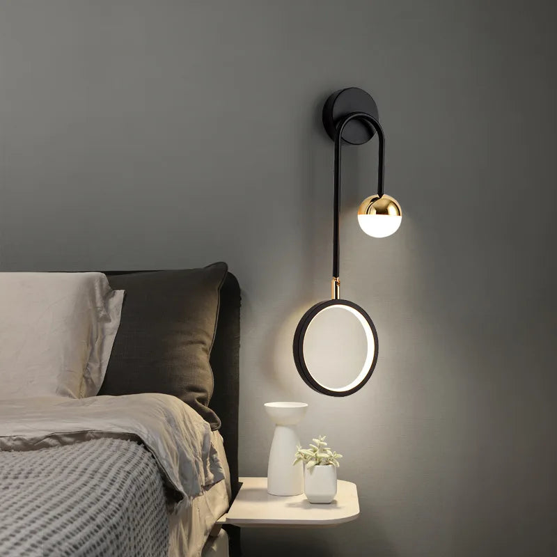 A stylish Hanging Circle Wall Light with a sleek black metal arm illuminates a bedside table adorned with two small vases and a plant, next to a bed featuring a gray headboard and neutral bedding. The modern design effortlessly incorporates geometric forms to elevate the space.