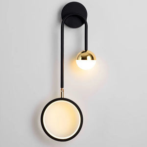 The Hanging Circle Wall Light is a modern wall sconce that features a minimalistic design with a black circular mounting base, gold half-sphere light fixture, and a circular illuminated ring hanging below. This stylish Nordic lamp showcases sleek geometric forms and elegance with its black metal arm.