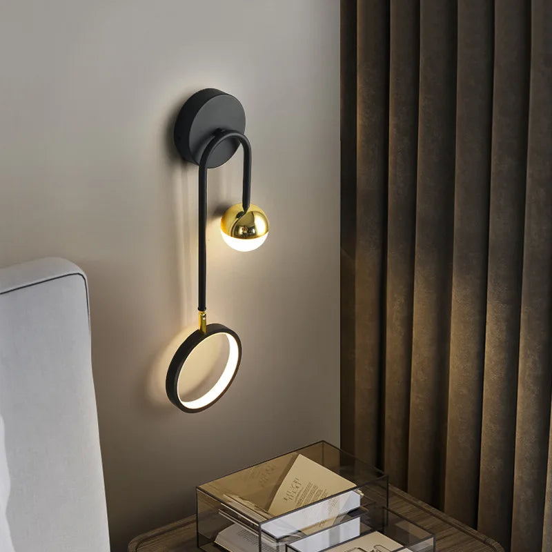 The Hanging Circle Wall Light, featuring a stylish Nordic design with a gold finish and black metal arm, illuminates the area next to a neatly made bed and side table adorned with books and a glass storage box, adding an elegant touch of geometric forms to the room.
