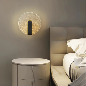 A contemporary Beautiful Wall Sconce emits warm light beside a sleek, grey nightstand and bed with white linens in a minimalist bedroom.