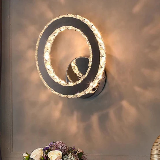 The Crystal Dark Frame Wall Sconce, featuring a sleek, modern circular design with two LED light elements, is mounted on the wall. A bouquet of flowers is partially visible at the bottom of the image.