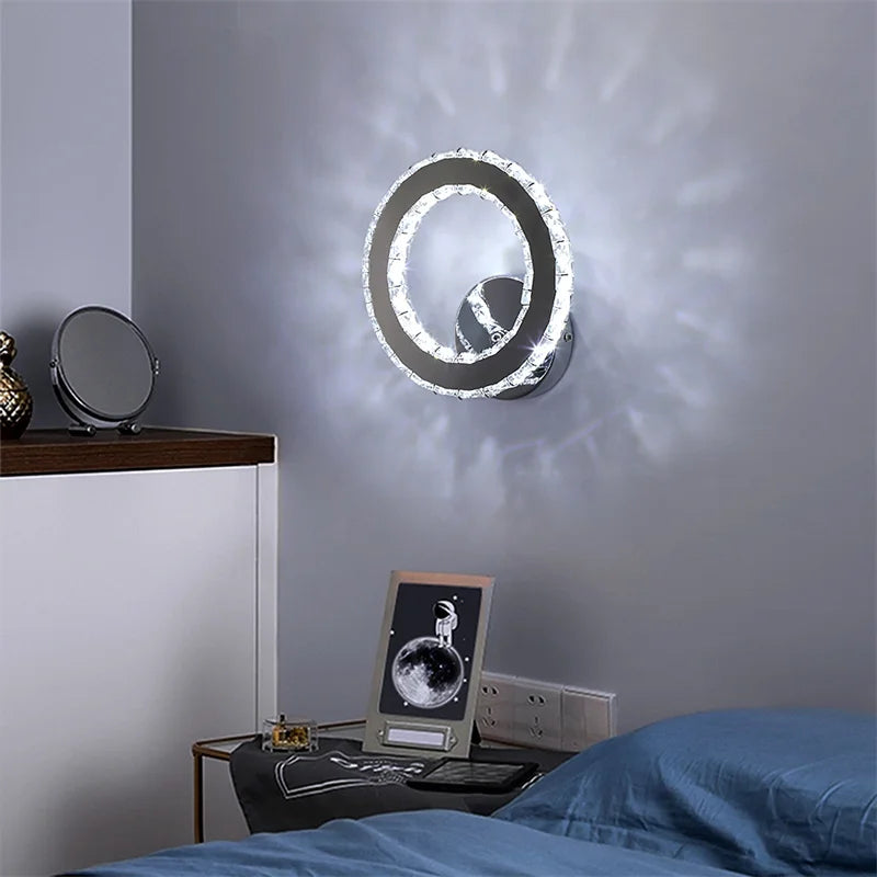 A Crystal Dark Frame Wall Sconce with a circular design is mounted above a nightstand adorned with a photo and cosmetic items. A neat blue bed is adjacent. The LED lights cast a radiant pattern on the gray wall.
