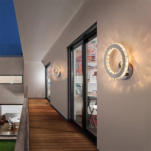 The Crystal Dark Frame Wall Sconce, with its sleek, circular design, illuminates a balcony featuring wood flooring and adjacent floor-to-ceiling windows. The interior appears to be a residential space. As the LED lights glow softly, the sky transitions to dusk.