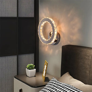 The Crystal Dark Frame Wall Sconce, featuring a modern circular design with two concentric rings and LED lights, illuminates the bedroom with a warm glow. On the bedside table, there's a potted plant and a photo frame next to the bed adorned with a patterned pillow.