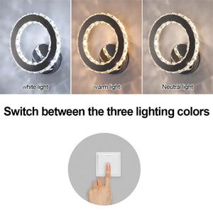 The Crystal Dark Frame Wall Sconce offers three lighting color options: white, warm, and neutral. Below, a finger presses a switch to change the LED lights. Text reads, "Switch between the three lighting colors.