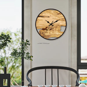 A Wood Circle Wall Clock featuring black hands and Roman numerals is mounted on a white wall. Below it, a black chair is positioned next to a table decorated with small white cups and a plant, adding charm to the setting.