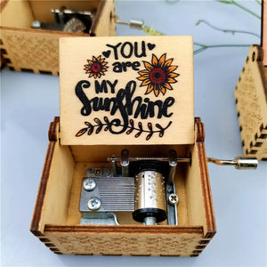 The Music Box - You Are My Sunshine features a romantic rotary handle and an open lid that reveals the engraved message "You are my sunshine" along with two sunflowers. The visible internal mechanism of this charming wooden music box makes it a delightful keepsake.