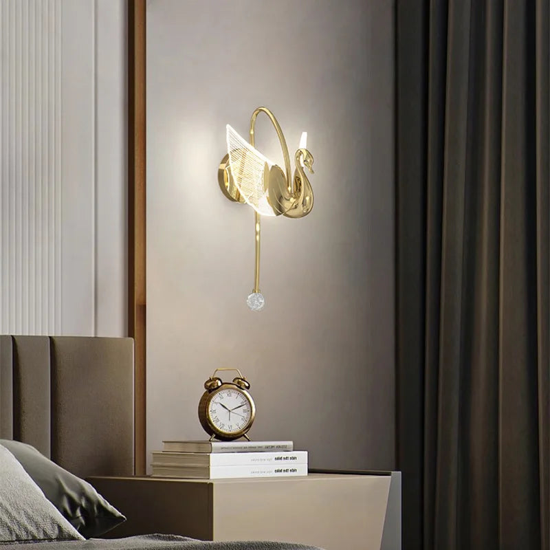 A lit Swan Wall Light, featuring a glass pendant, hangs elegantly above a bedside table decorated with an alarm clock and a stack of books in a modern bedroom.
