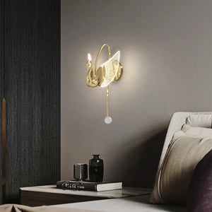 The Swan Wall Light, a wall-mounted decorative swan sconce, illuminates the bedroom above a nightstand filled with books and decorative items. Its gracefully curving wings and neck emit a soft glow, enhancing the elegant indoor lighting.