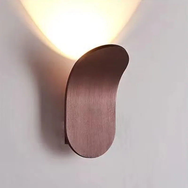 The Curved Wall Light, a Nordic-inspired wall lamp featuring a brushed metal design in brown, emits light upwards and is mounted on a white wall. Perfect for living spaces seeking modern light fixtures that seamlessly blend elegance and functionality.