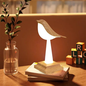 A Bird Table Night Light, crafted with elegant wooden design, perches gracefully on top of a stack of books on a table. It's placed next to a vase containing a plant and several lettered blocks. This distinctive bird-shaped lamp introduces a contemporary flair to any room.