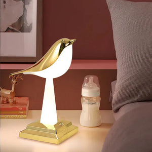 A Bird Table Night Light in a minimalist gold bird shape sits on a bedside table next to a baby bottle and children's book.