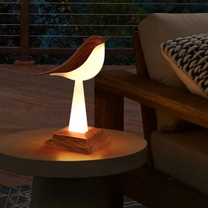 The Bird Table Night Light, crafted with elegant craftsmanship and a wooden base, emits a soft glow on a round table next to a couch with a knit pillow on a deck at dusk.