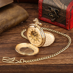 A Golden Roman Pocket Watch with an open cover and visible chain rests on a wooden surface beside a leather-bound book and a small decorative chest.